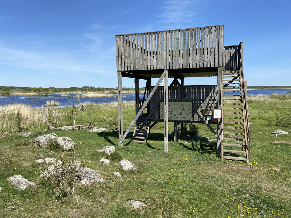 Ett fågeltorn i två våningar står på en gräsyta med flera stenblock. I bakgrunden syns sjön.