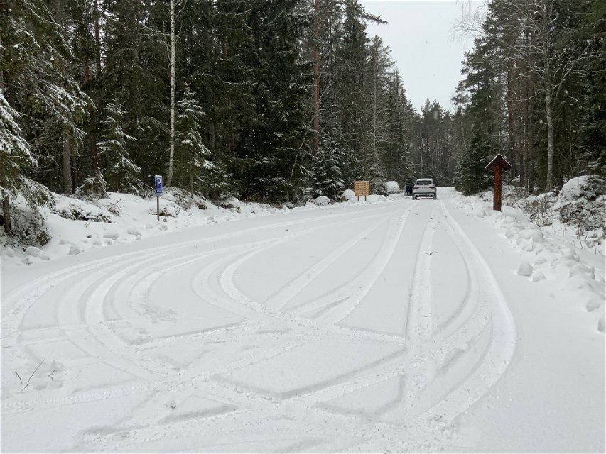 En snöig parkeringsplats längs en skogsbilväg. En informationstavla och en stigvisare står på sidan av vägen. Längst bort på vägen står en bil.