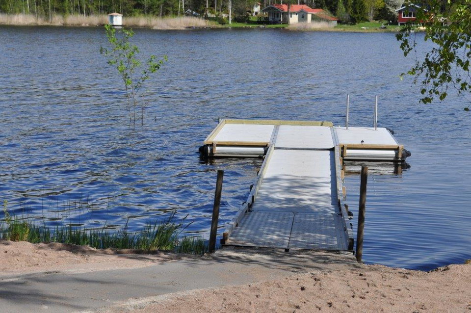 En bred ramp leder ner till en flytbrygga med skyddskant runtom. Från bryggan leder en stege ner till vattnet.