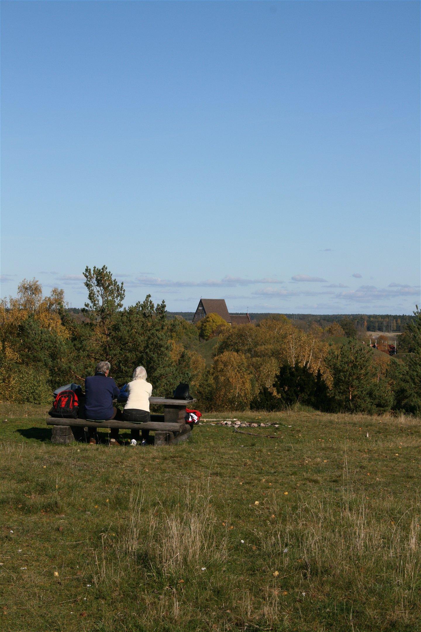 Ett äldre par sitter vid ett bänkbord uppe på en höjd. De blickar ut över träd och en högre byggnad i bakgrunden.