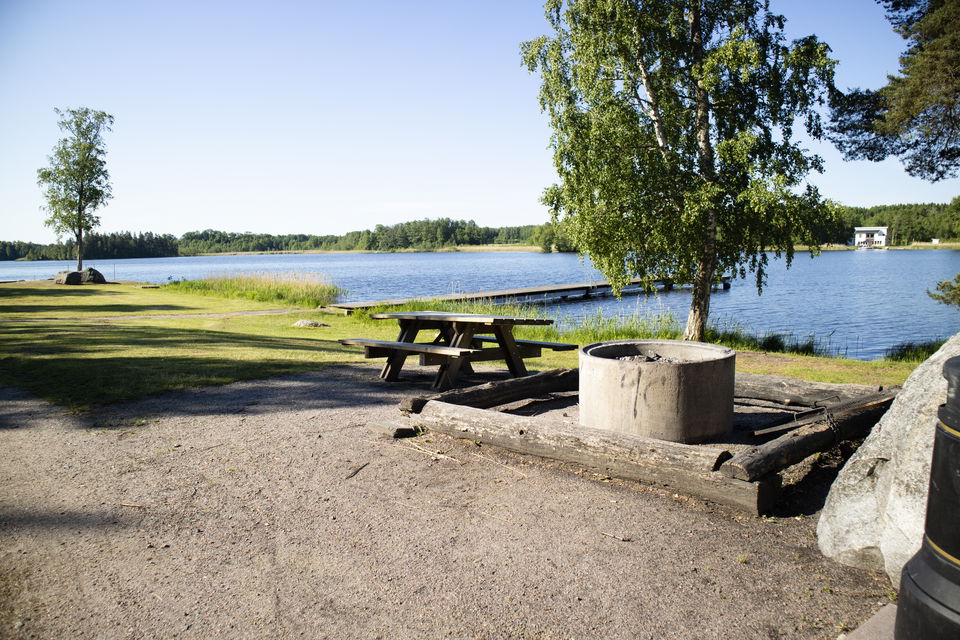Grillplats med bänk och stockar att sitta runt, träd och sjö i bakgrunden.