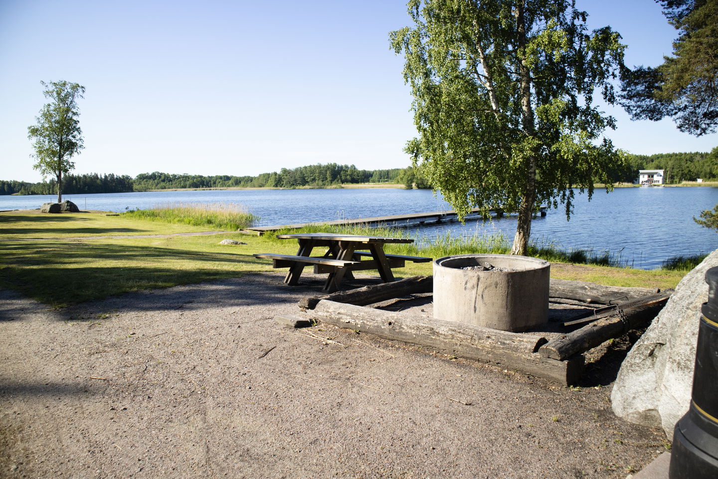 Grillplats med bänk och stockar att sitta runt, träd och sjö i bakgrunden.