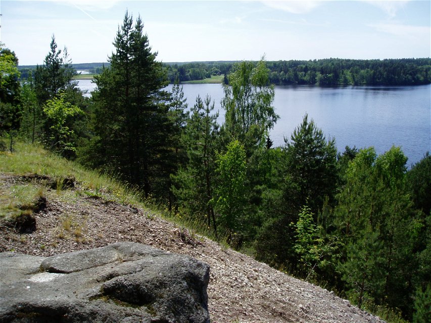 Utsikt över sjön uppe på höjd på en ås. Det sluttar mycket brant nedåt och i förgrunden står en stor sten.
