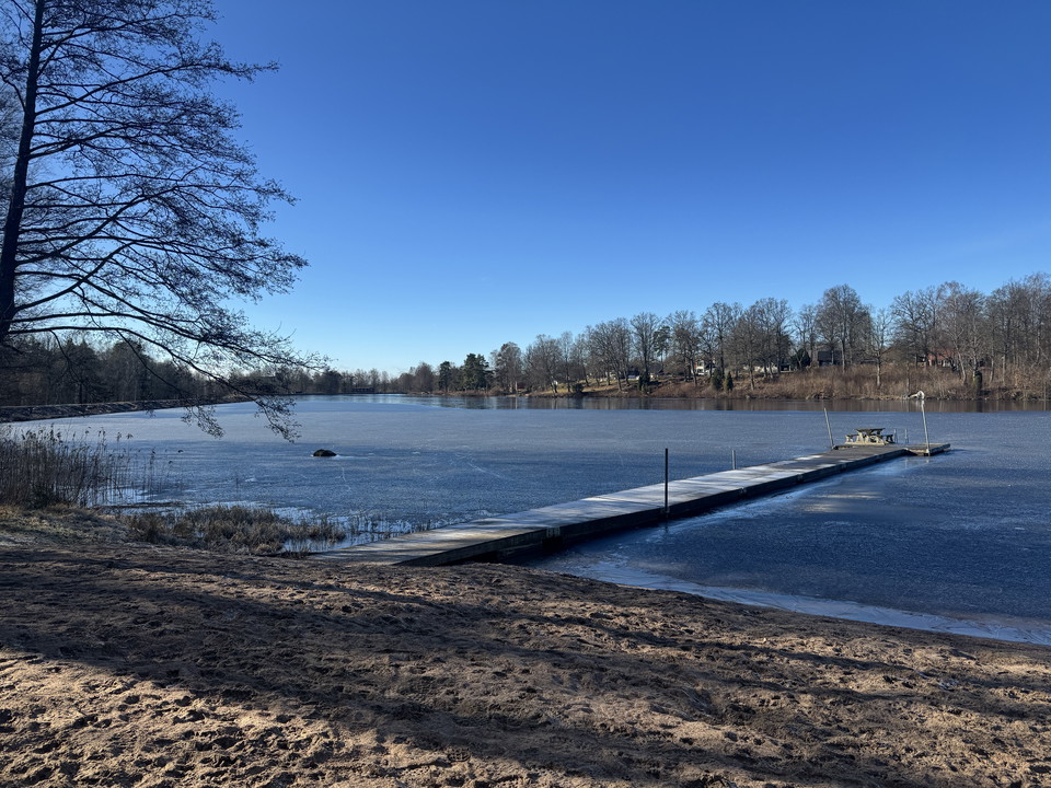 En smal sandstrand med en brygga ut i Helge å. Träden runt omkring är kala och det är lite is på vattnet.