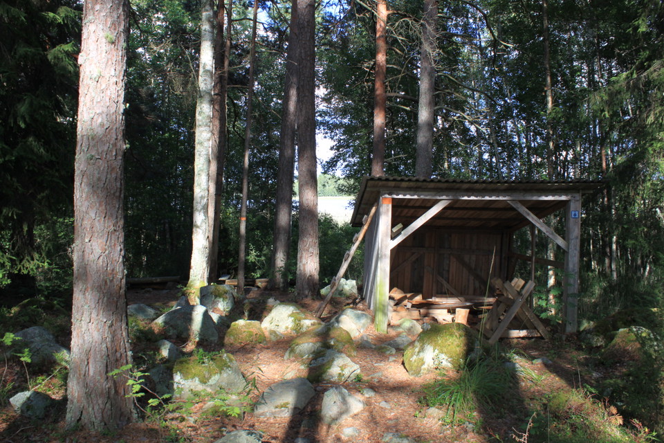 Ett vedförråd står på ojämn och stenig mark i en skogsglänta.
