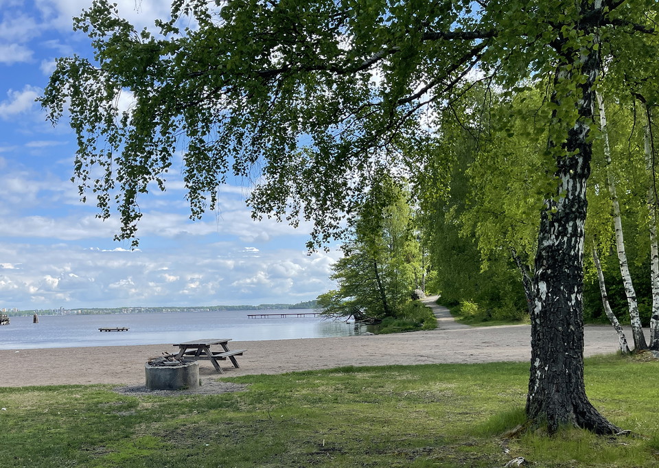 Badplats med sandstrand. En grillstad och ett picknickbord. På avstånd en grusad stig och en brygga. 