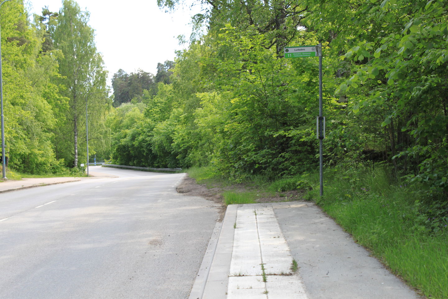 Vid en bilväg finns en busshållplats markerad med en skylt. På båda sidorna av vägen är det skog.