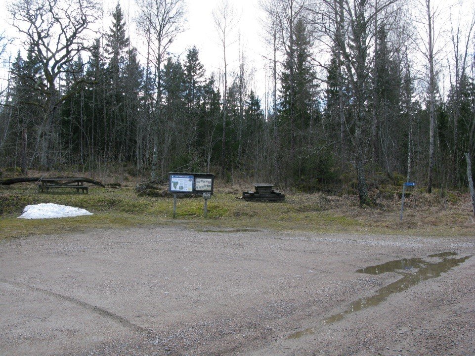 En grusad parkeringsplats med en rastplats vid ena sidan. På rastplatsen står två bänkbord och en informationstavla på något ojämn mark.