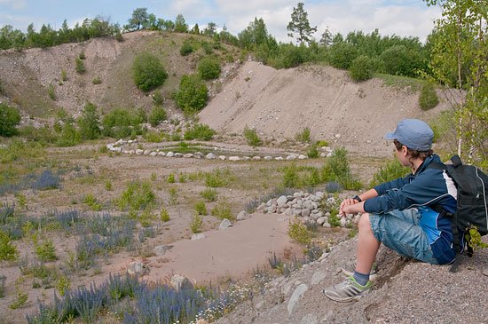 En person sitter uppe på en grushös och blickar mot en damm. Dammen ligger i botten av en djup grusgrop.