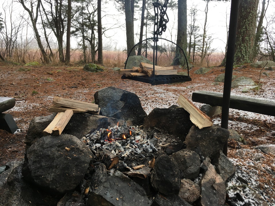 Över en eldstad med stenar runtom hänger ett grillgaller. Grillplatsen står i en glänta med lite snö på marken och bredvid finns sittbänkar. 