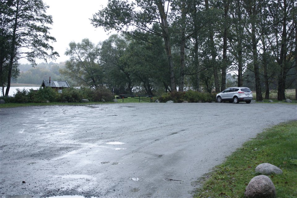 En bil står på en rymlig parkering med träd vid ena kanten. Från parkeringen ser man vatten och det finns flera vattenfyllda gropar på marken.