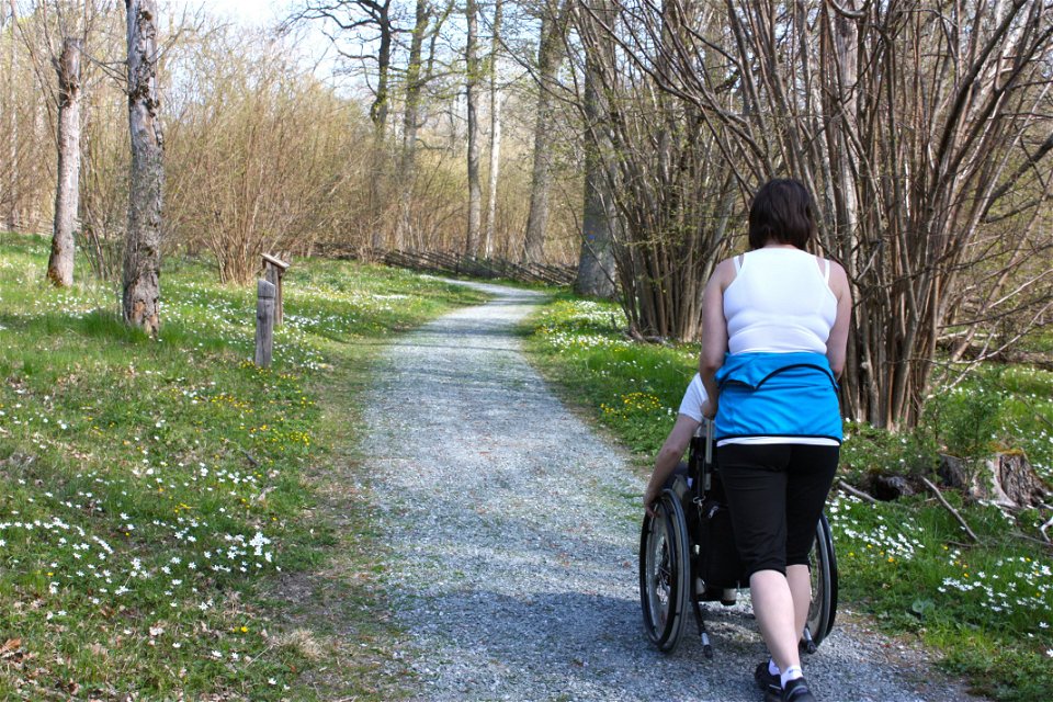 En kvinna skjutsar en person i rullstol längs en grusad promenadstig. Det är gott om blommor och träd runtom.