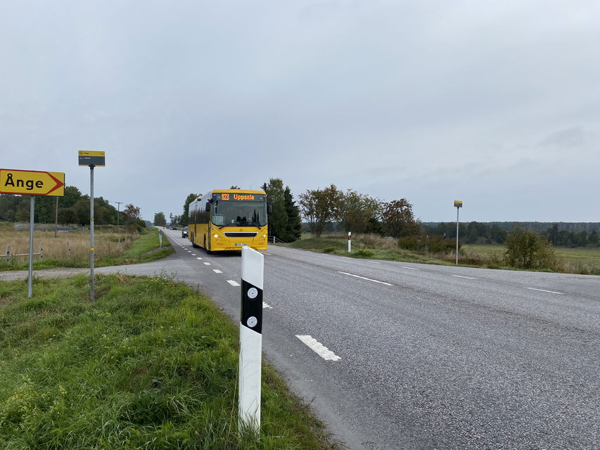 Asfalterad väg med gräs på sidan. Kommer en gul UL-buss. Gul skylt pekar höger, text Ånge