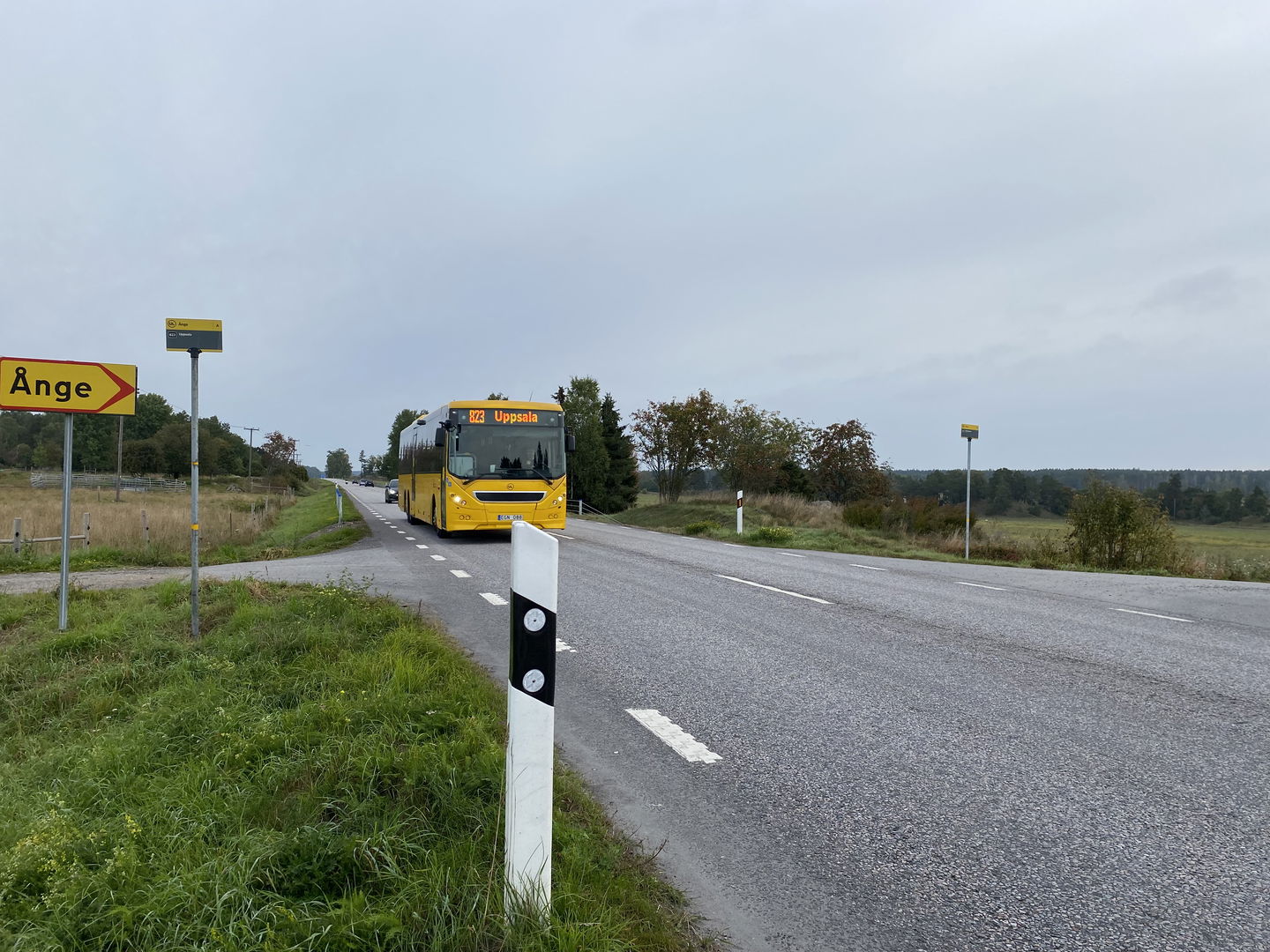 Asfalterad väg med gräs på sidan. Kommer en gul UL-buss. Gul skylt pekar höger, text Ånge