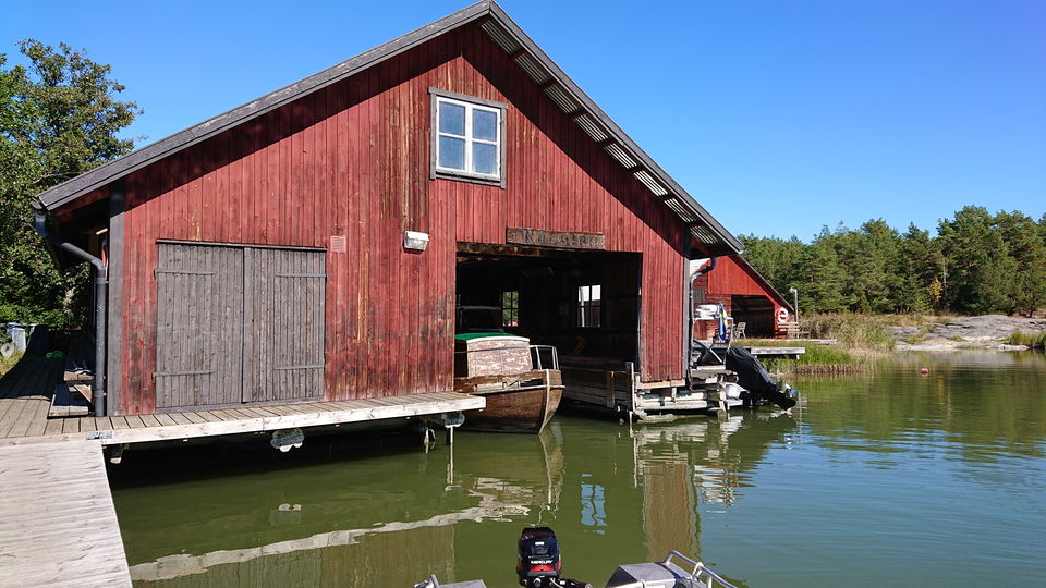 Ett båthus med en båt förtöjd inuti. Utanför huset finns en brygga och i bakgrunden syns ett annat båthus.