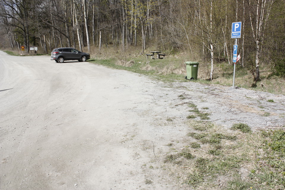 En bil står på en avlång grusad parkeringsplats. Framför parkeringen finns en skogsdunge med rastplats, soptunna och parkeringsskylt.