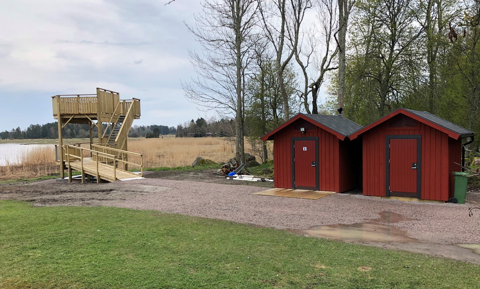 Tillgängligt fågeltorn, liten vattenspegel och vass i bakgrunden. Två små röda byggnader, hårdgjord yta framför