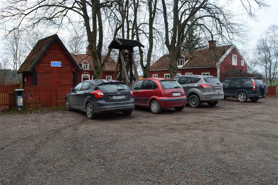 Fyra bilar står bredvid varandra på en grusad parkeringsplats. Framför bilarna står en gård med flera träbyggnader.