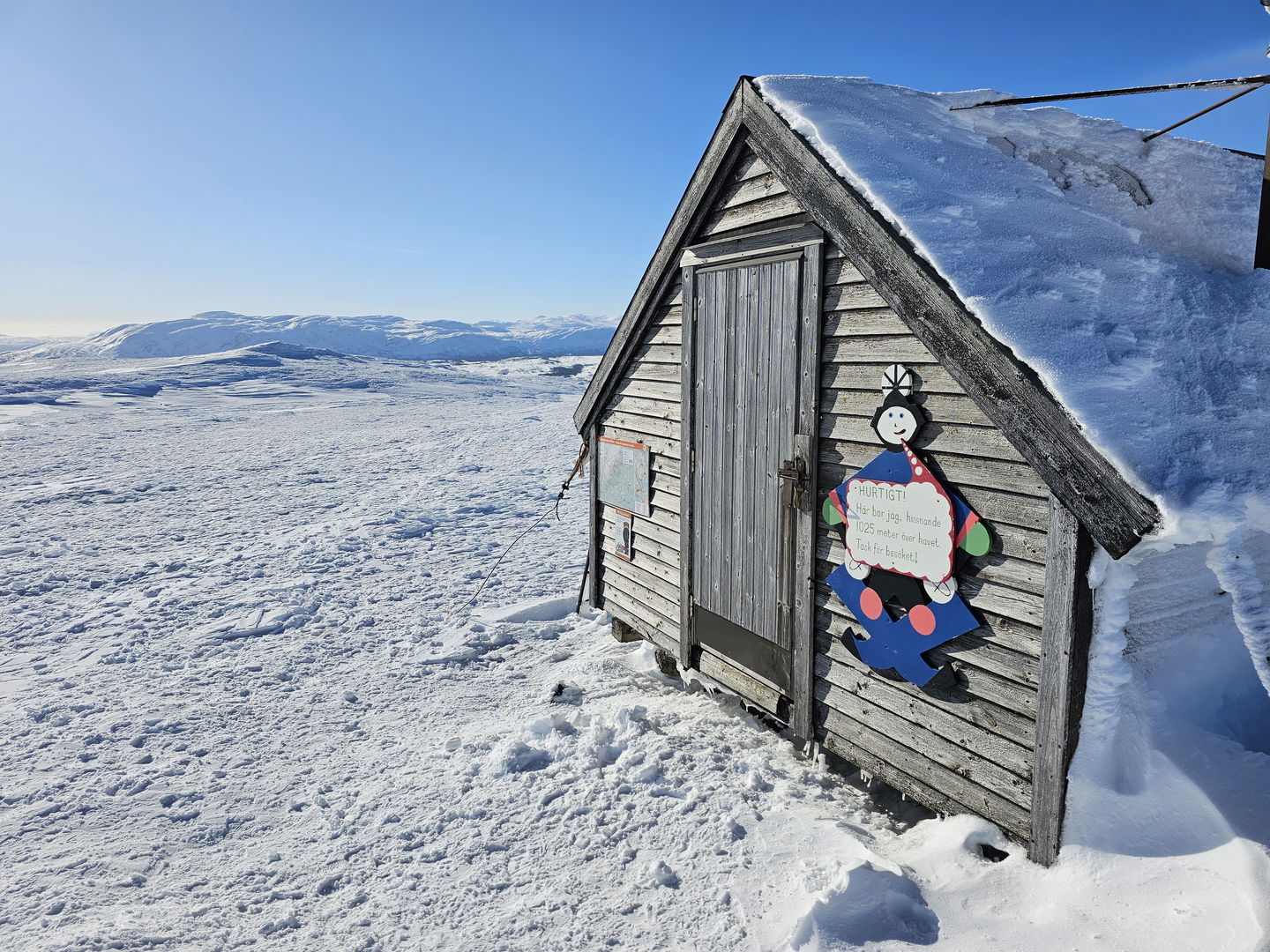 Fasaden av en liten grå stuga på toppen av ett snöigt fjäll