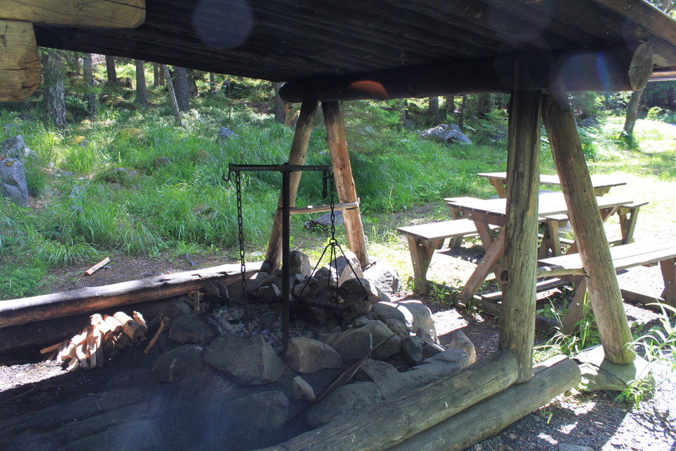 Närbild på en eldstad med stenar runtom och ett grillgaller över. Eldstaden står under tak och två bänkbord står utanför.