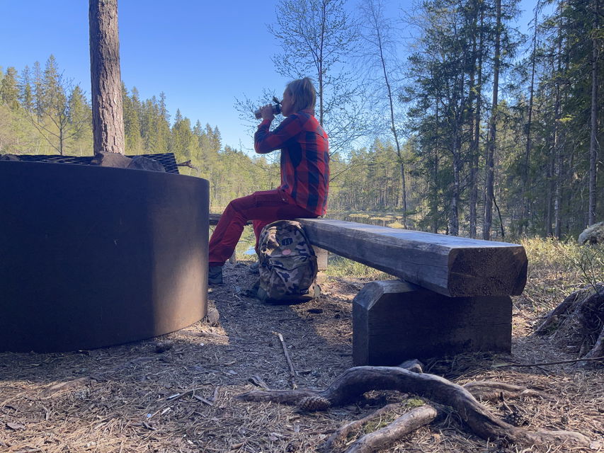 En rödklädd person sitter på en bänk och dricker ur en termos.