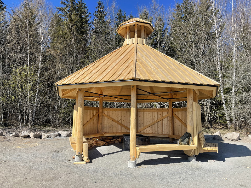 Åttakantig träbyggnad där fem väggar är täckta. I byggnaden finns väggfasta bänkar och i mitten en grillstad. 