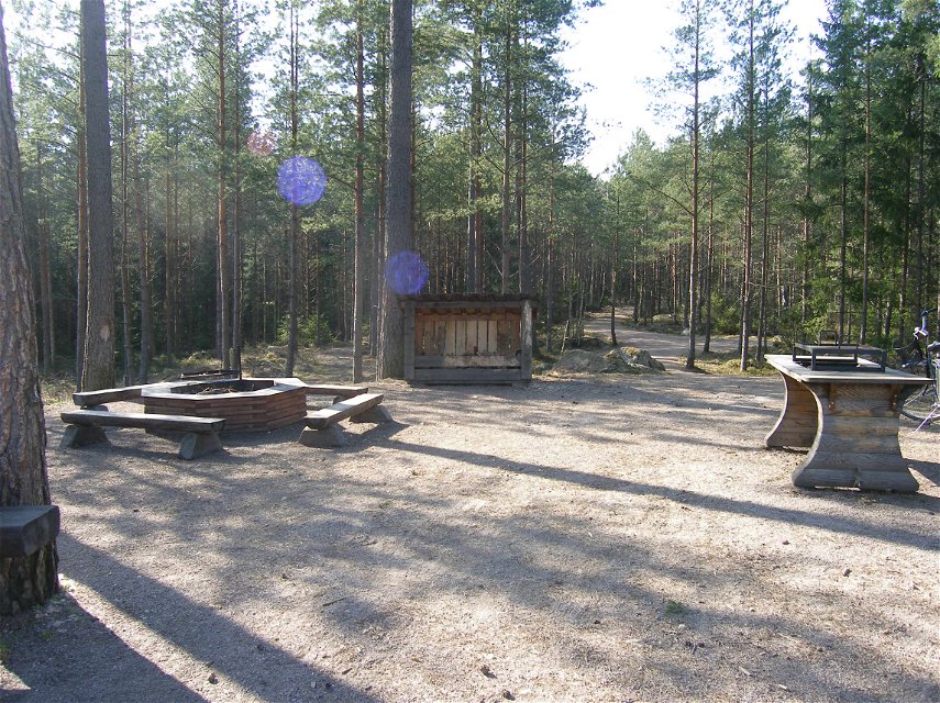 På en öppen grusad yta i skogen finns en grillplats med sittbänkar runtom och ett grillbord. I bakgrunden står en vedbod.
