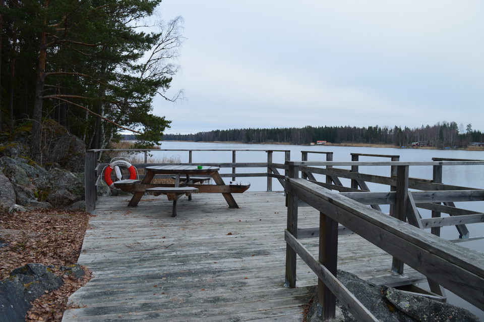 En brygga med handräcke leder fram till ett trädäck med ett bänkbord.