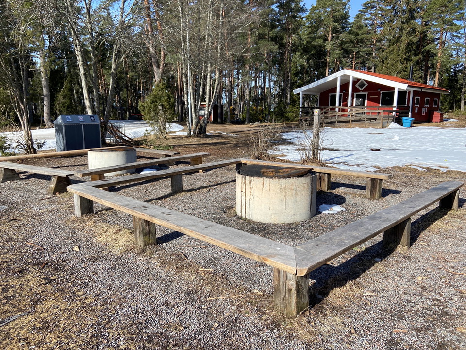 En eldstad i form av ett cementrör med sittbänkar i en fyrkant runt. I bakgrunden ses en röd stuga.