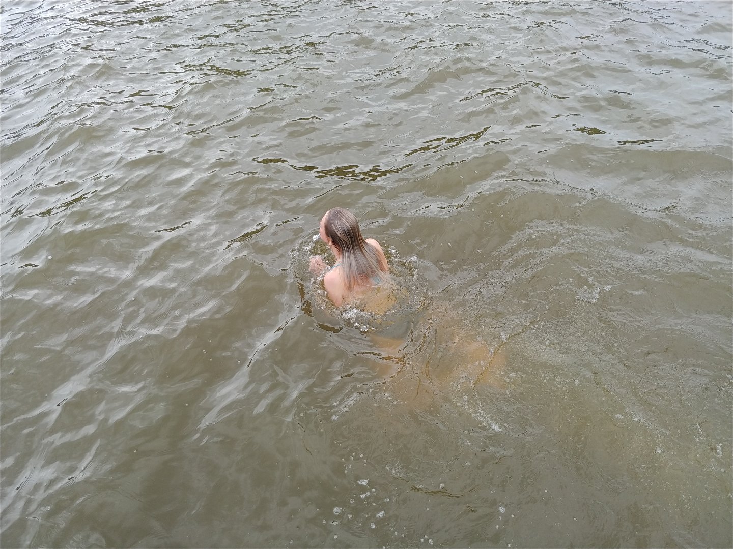 Person med brunt, långt hår som simmar i sjövatten, ansiktet är vänt från kameran.