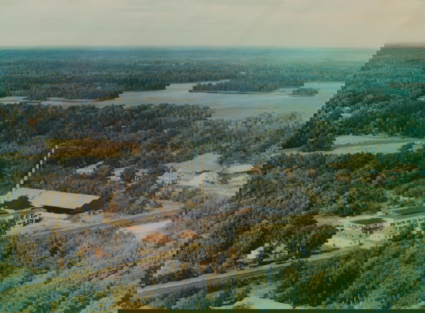 En långsmal fabriksbyggnad intill ett järnvägsspår är omringat av barrskog.