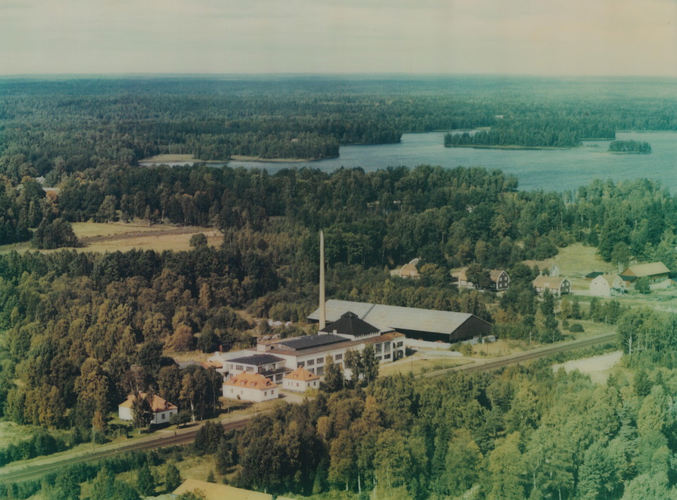 En långsmal fabriksbyggnad intill ett järnvägsspår är omringat av barrskog.