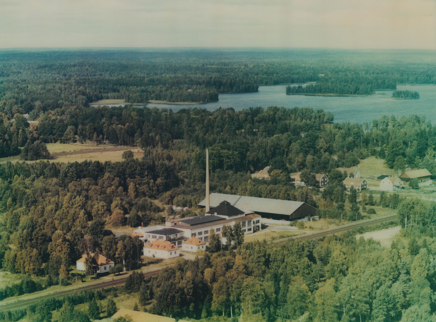 En långsmal fabriksbyggnad intill ett järnvägsspår är omringat av barrskog.