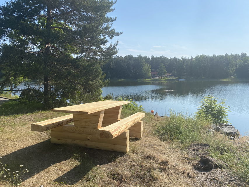 Ett träbord med fasta bänkar står på en gräsmatta. Vatten syns i bakgrunden