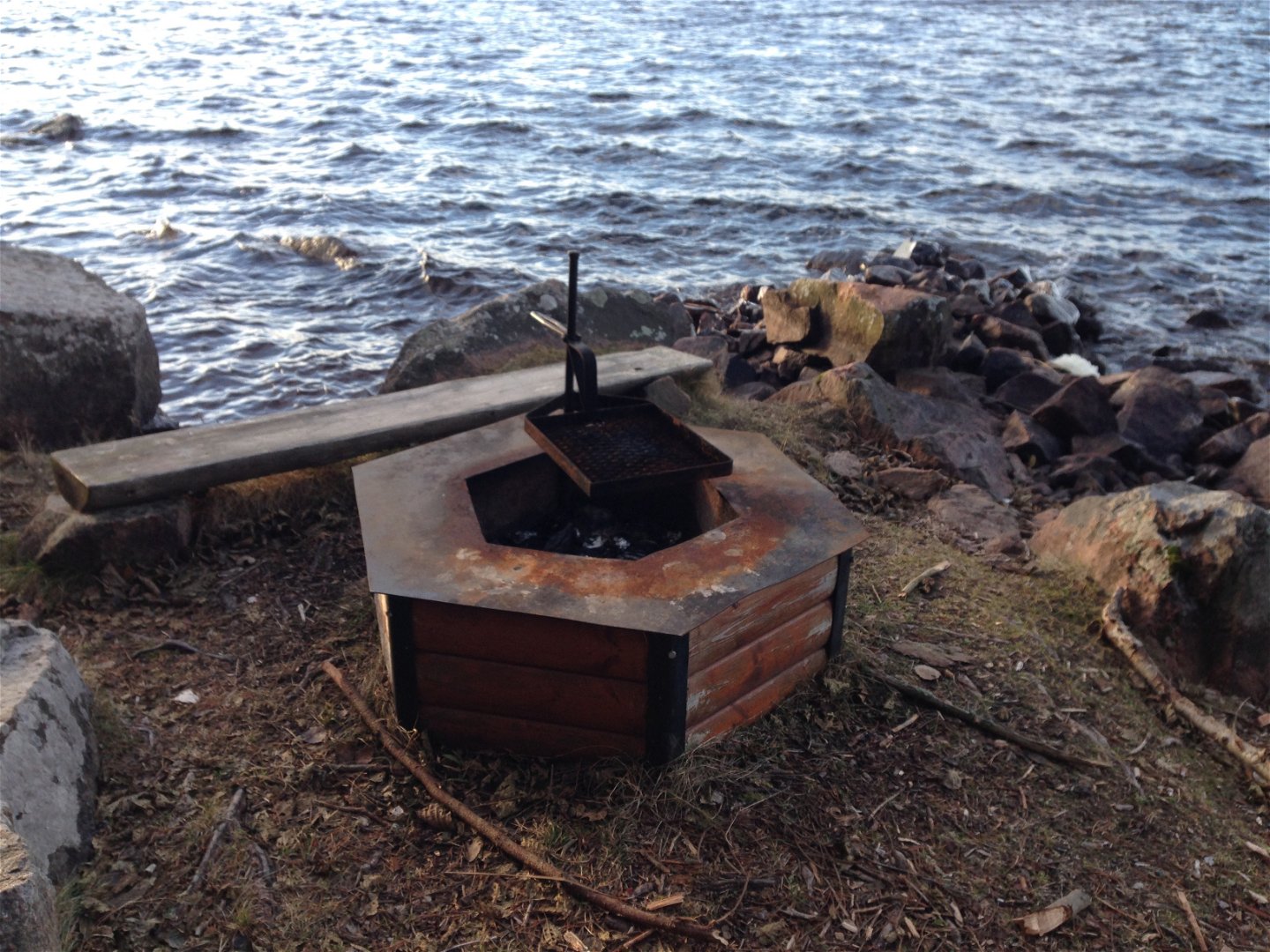 En grillplats med grillgaller och en låg sittbänk bredvid står vid en sjö.