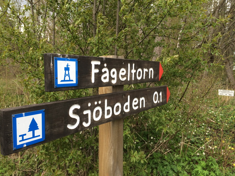 En hög stolpe står i skogen. På stolpen finns två skyltar som pekar åt höger med texten fågeltorn och sjöboden.