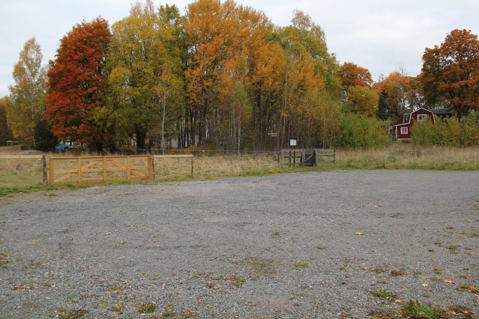 En rymlig, grusad parkeringsplats framför staketet in till en hage. Vid parkeringen finns en informationstavla och i bakgrunden skymtar ett hus fram mellan träd.