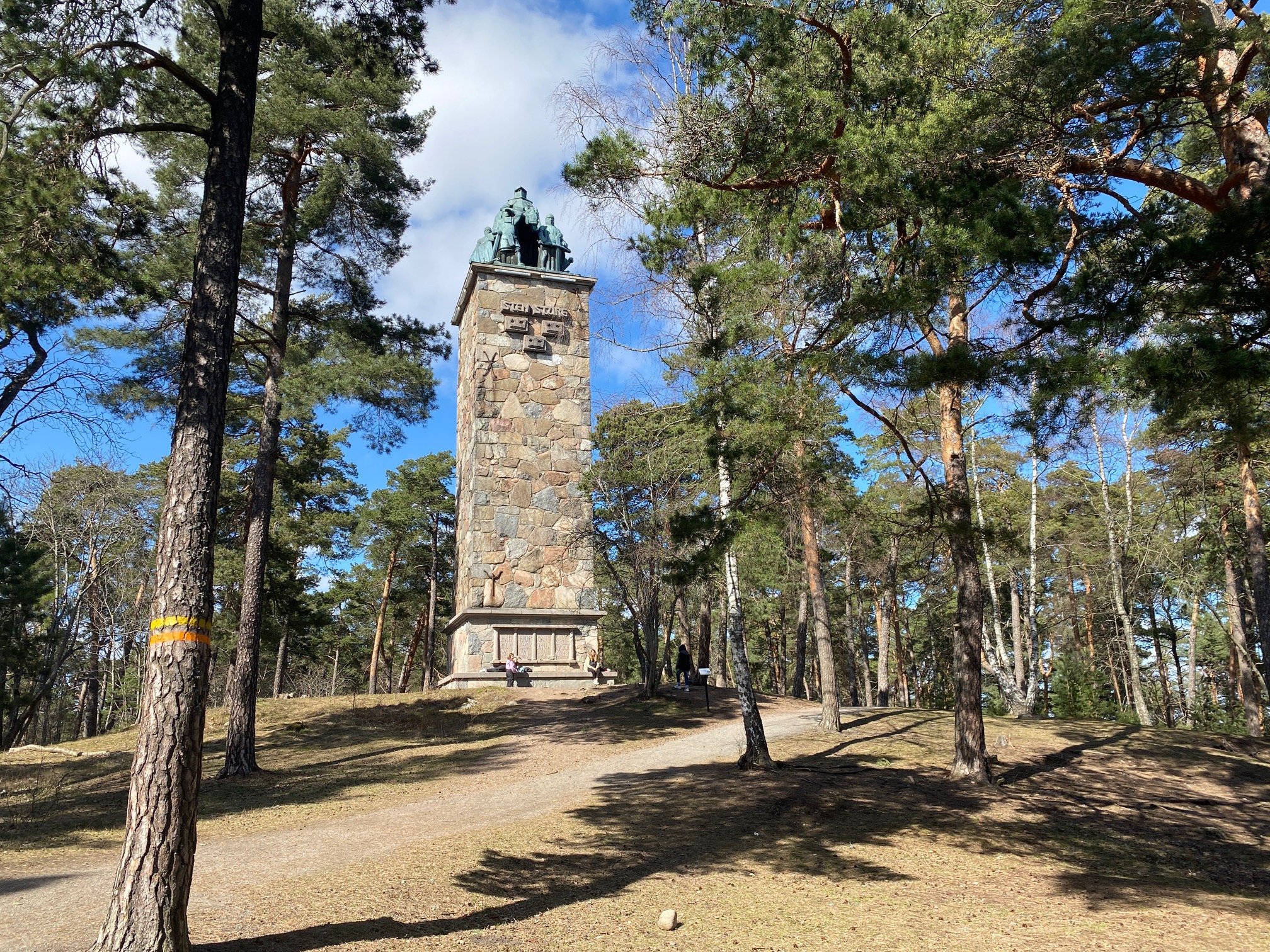 I en skog står ett högt torn av sten med en skulptur på toppen. På ena trädet finns en orange markering.