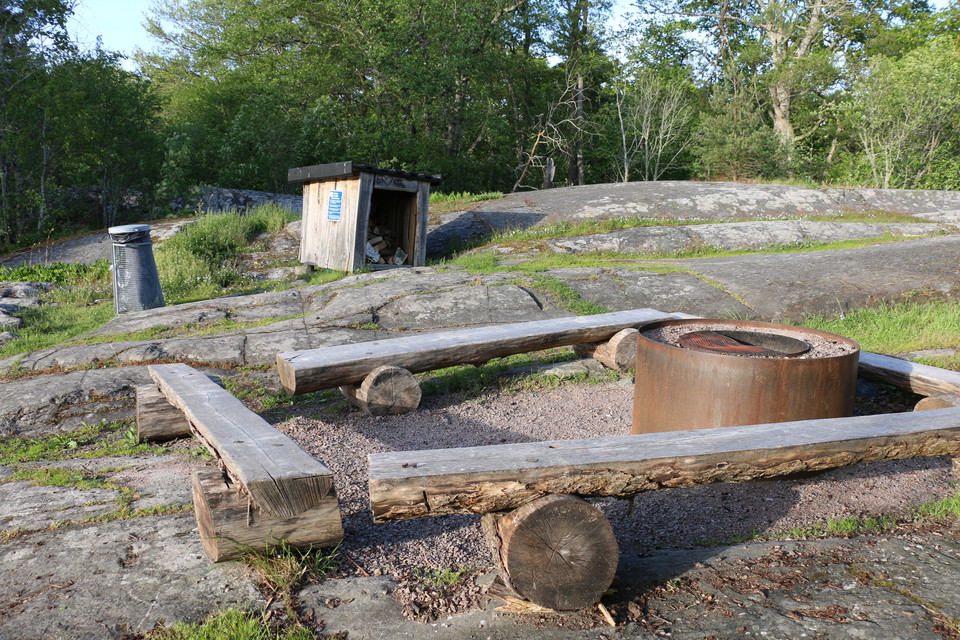 Eldstad med bänkar runtikring. I bakgrunden syns en soptunna och ett vedförråd.