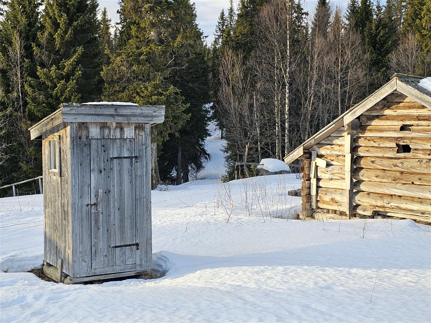 En liten träbyggnad som är ett utedass samt gavel på ett timrat hus. på marken ligger snö