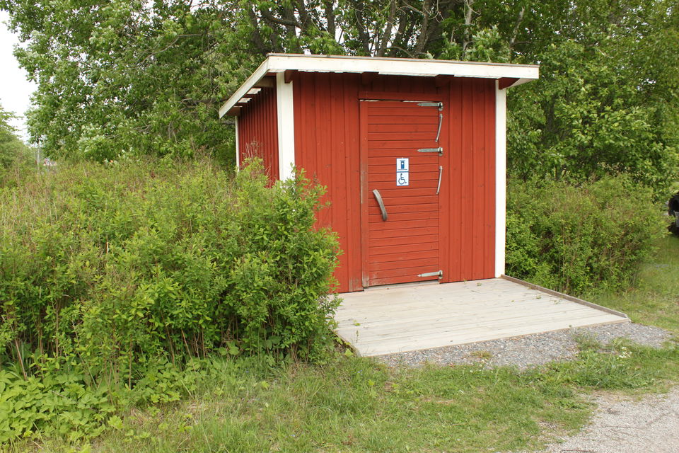 En bred träramp leder upp till en torrtoalett. Runtom toaletten är det buskage.