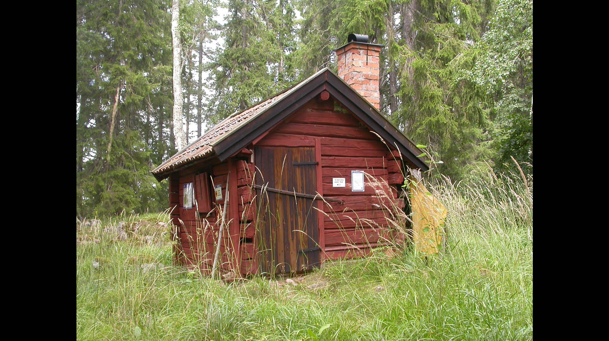 I en skogsglänta står en liten stuga av äldre modell. Huset har en skorsten.