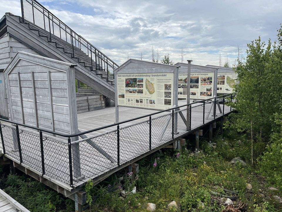 Fyra informationstavlor på rad vid en grånande träbyggnad.