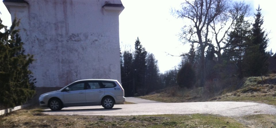 På en något lutande asfalterad parkeringsplats står en bil. I bakgrunden står Järnmagasinet.