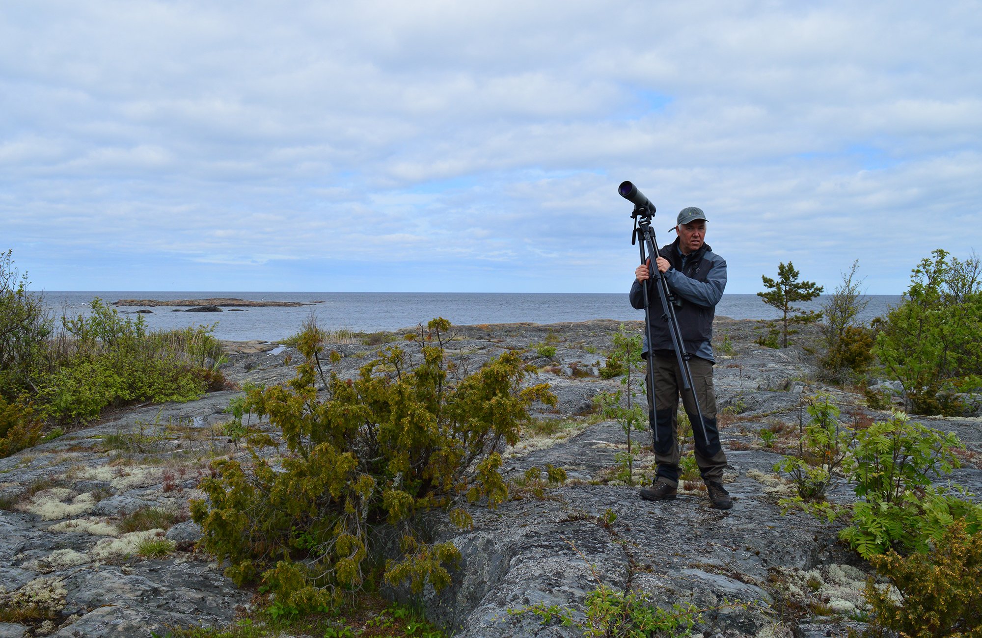 En man står på en stenhäll och håller i en tubkikare. I bakgrunden är det hav.