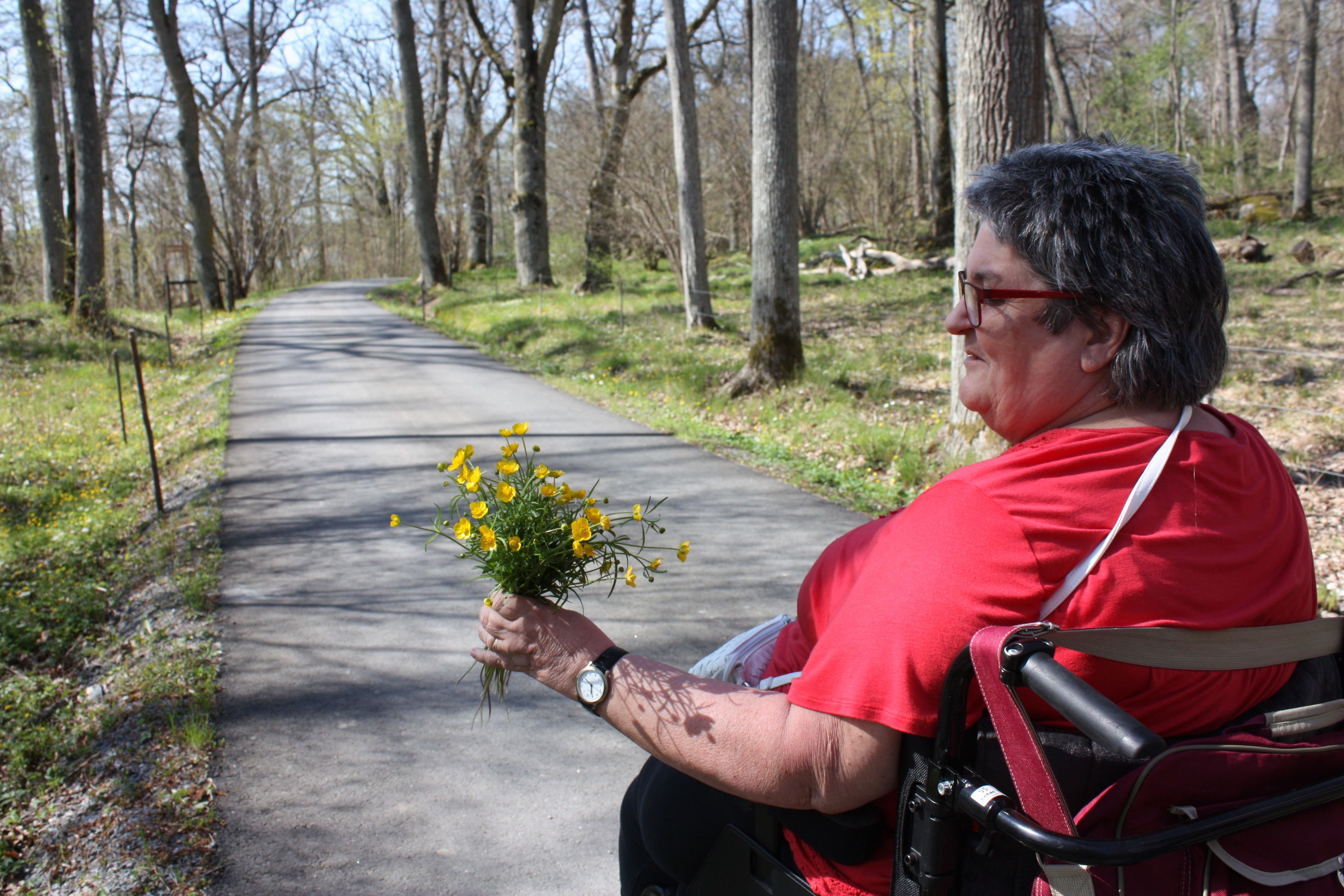 En kvinna håller i en bukett med gula blommor ute i en park. Kvinnan sitter i rullstol som står på en asfaleterad väg.