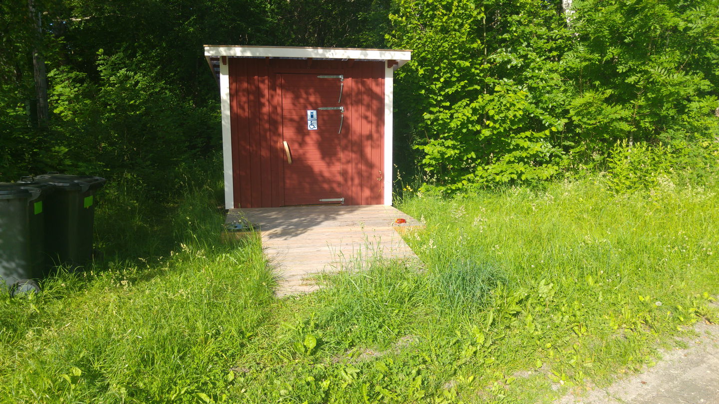 En bred ramp leder upp till en torrtoalett som står på gräs. Bakom toaletten står många träd.