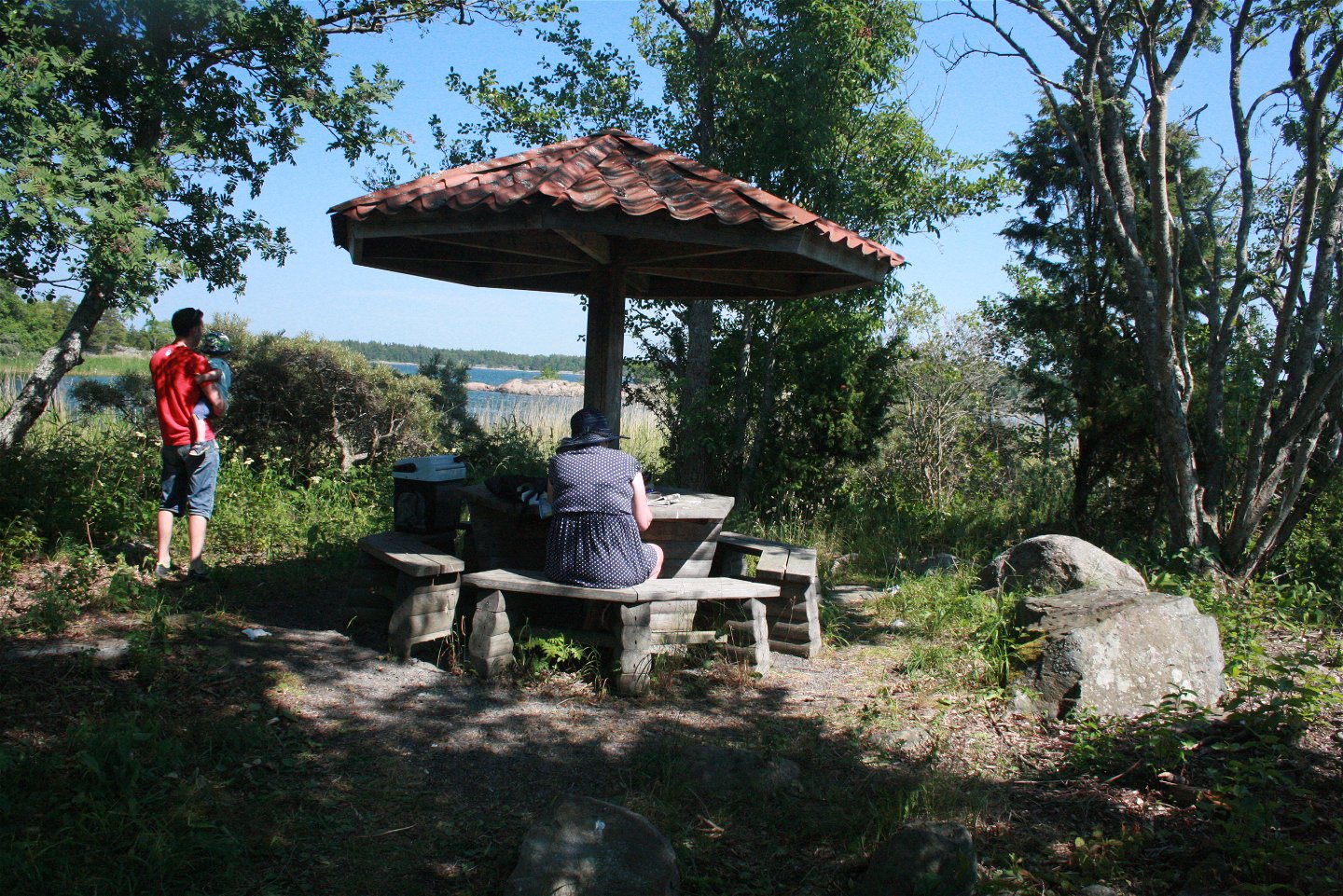 I en skogsglänta står ett runt bord med en parasoll mitt i och bänkar runtom. En kvinna sitter vid bordet och en man med ett barn i famnen står bredvid.
