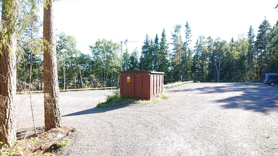 En stor grusad parkeringsplats med ett stort elskåp i mitten. Runtom parkeringen är det skog. 