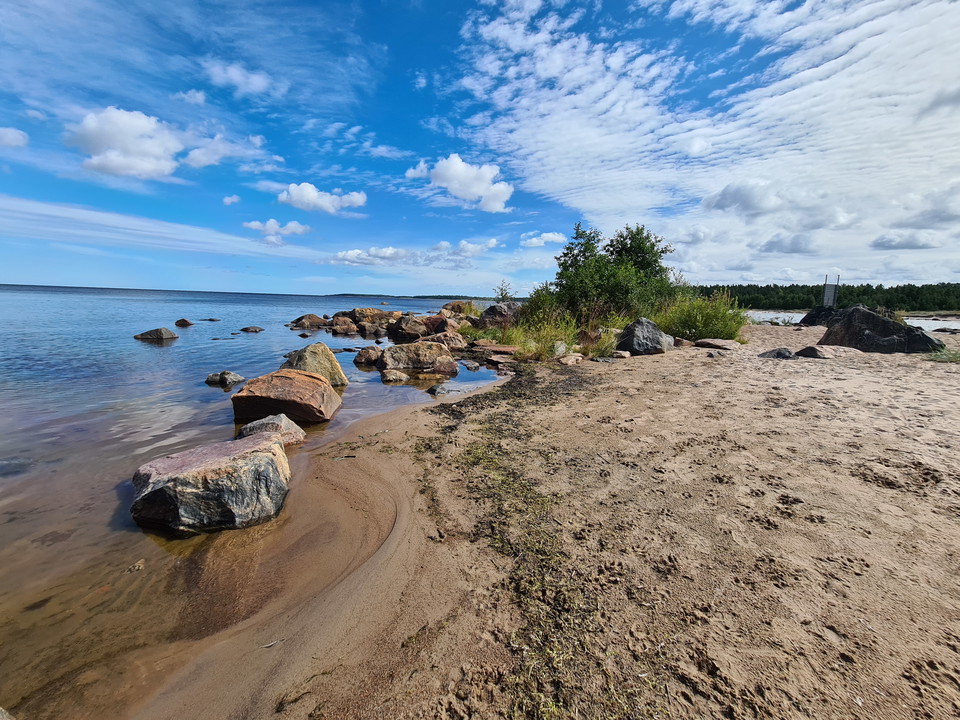 Udde i havet med sandstrand. I vattenbrynet ligger några stora stenar. Några buskar växer på sandstranden.
